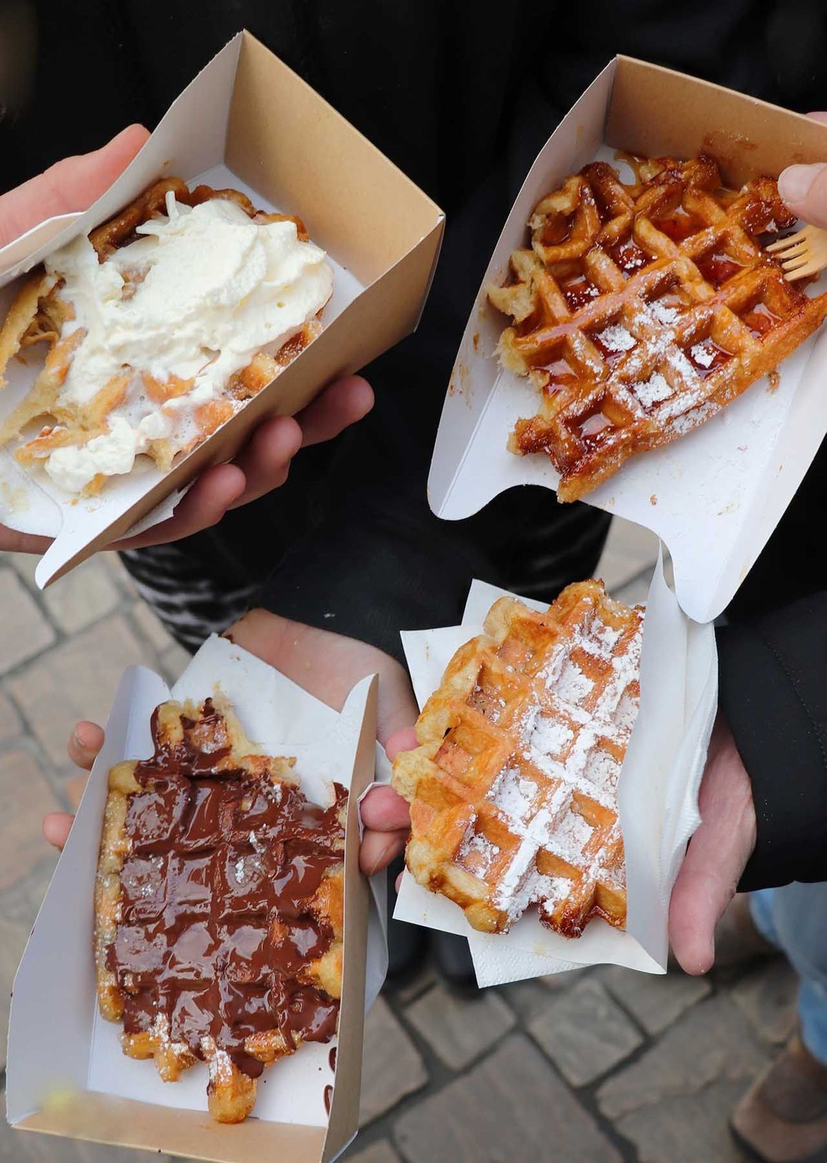 Four hands holding fresh Belgian waffles topped with cream, chocolate and dusted sugar at a street stall